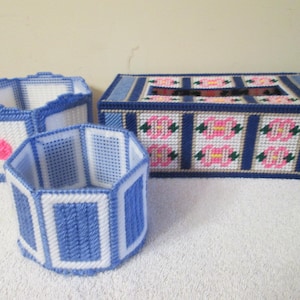 Peut inclure: Trois articles faits à la main en canevas plastique : deux petits contenants et un cache-boîte à mouchoirs. Les contenants sont bleus et blancs avec un accent rose. Le cache-boîte à mouchoirs est bleu avec des motifs floraux roses et verts.