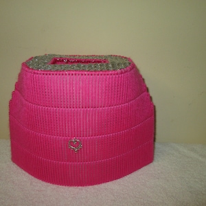 May include: A pink tissue box cover with a silver heart design. The cover is made of plastic and has a woven texture.