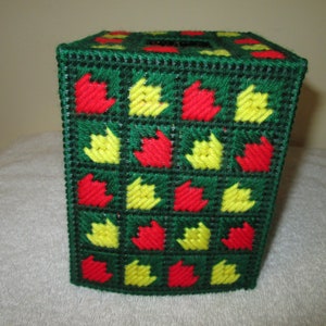 Può includere: Copertura per scatola di fazzoletti fatta a mano di forma quadrata. La copertura è realizzata in tela di plastica verde con un motivo di forme rosse e gialle a forma di foglia. La parte superiore ha un'apertura quadrata.