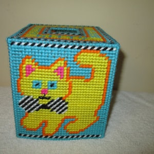 Op de afbeelding: Een vierkante tissueboxhoes gemaakt van plastic canvas. De hoes is turquoise met een gele kattenontwerp omlijnd in oranje. De kat heeft roze oren en blauwe ogen, en een zwart-wit gestreepte vlinderdas.