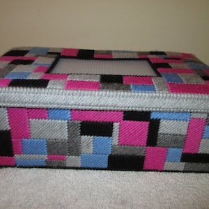 May include: Rectangular box with a lid, featuring a patchwork design in pink, black, gray, and blue squares. The lid has a rectangular opening and is trimmed with silver-colored piping. The box sits on a white surface.