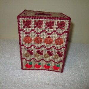 May include: A red, white, and brown crocheted tissue box cover with a fall theme. The cover features pumpkins, leaves, and apples.