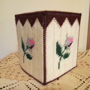 May include: A white tissue box cover with a brown border and embroidered pink and white roses. The cover is made of plastic canvas and has a woven texture.