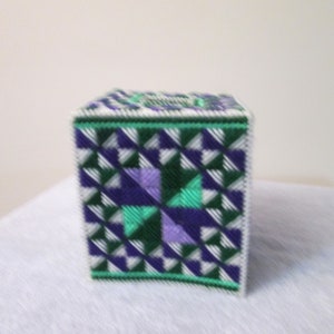 May include: A square tissue box cover made of plastic canvas with a geometric pattern in shades of green, purple, and white.