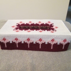 May include: A white tissue box cover with a pink and burgundy floral design. The cover is made of plastic canvas and has a burgundy border.