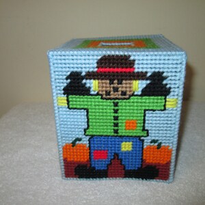 May include: A blue and white tissue box cover with a scarecrow design. The scarecrow is wearing a brown hat, green shirt, blue pants, and orange and yellow patches. The scarecrow is standing in front of two orange pumpkins.