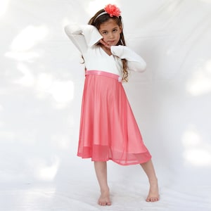 May include: A young girl wearing a white long-sleeved dress with a pink tulle skirt and a pink flower headband. She is standing on a white background.