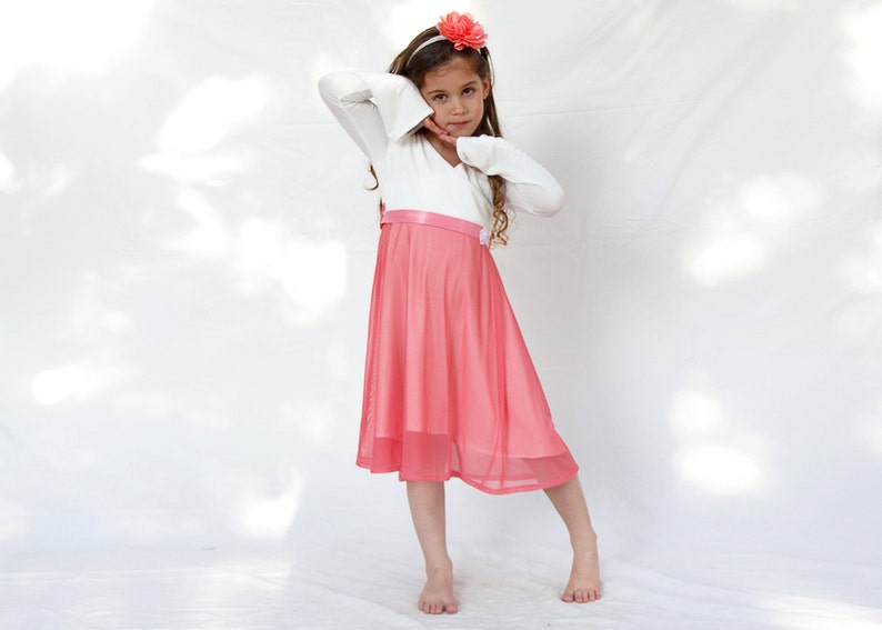 May include: A young girl wearing a white long-sleeved dress with a pink tulle skirt and a pink flower headband. She is standing on a white background.