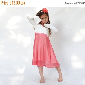 May include: A young girl wearing a white long-sleeved top with a pink tulle skirt and a pink flower headband.