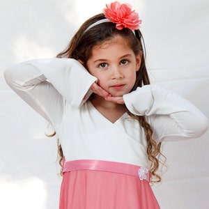 May include: A young girl with long brown hair wears a white long-sleeved dress with a pink skirt and a pink flower headband.