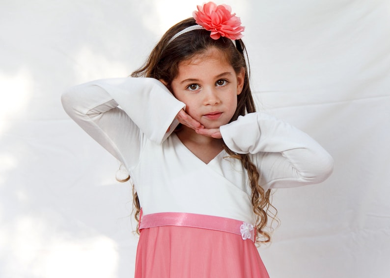 May include: A young girl with long brown hair wears a white long-sleeved dress with a pink skirt and a pink flower headband.