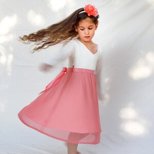 May include: A young girl wearing a white long-sleeved top with a pink skirt and a pink flower headband. The skirt has a white waistband and a pink bow.