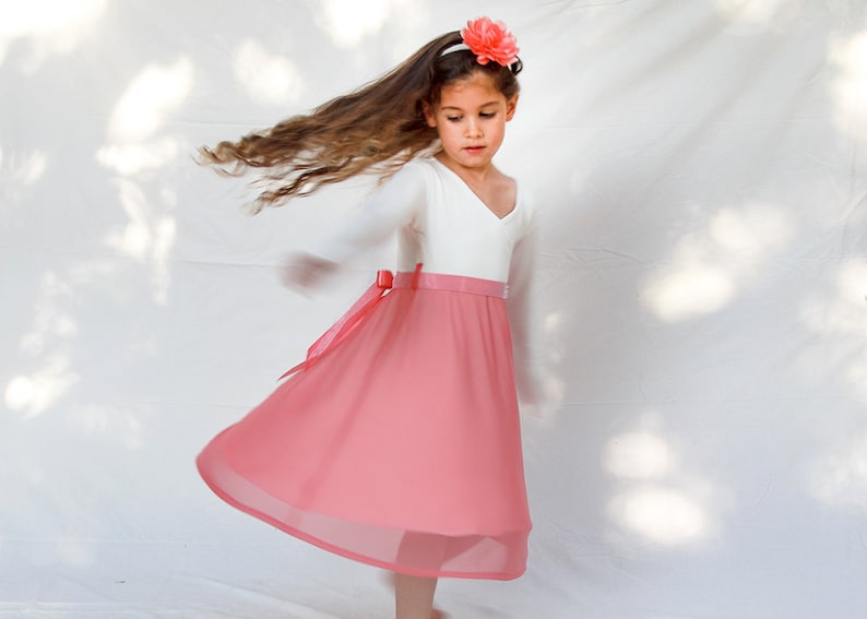 May include: A young girl wearing a white long-sleeved top with a pink skirt and a pink flower headband. The skirt has a white waistband and a pink bow.