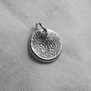 May include: A small, circular silver pendant with a textured pattern. It features a loop for attaching to a chain. The pendant's surface is entirely covered in a detailed, textured design.