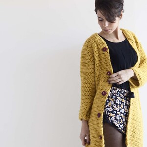 Puede incluir: Un cárdigan de ganchillo de color amarillo mostaza con mangas largas, que presenta un patrón de punto texturizado y cinco botones circulares de color morado oscuro en la parte delantera. Se lleva sobre una camiseta negra y pantalones cortos estampados.