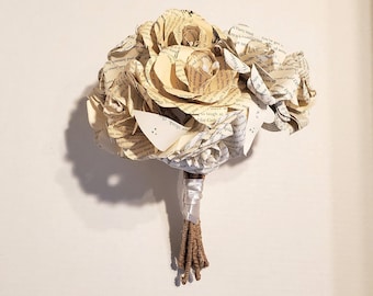 May include: A bouquet of paper flowers made from the pages of a book. The flowers are white and cream colored and are tied together with a white ribbon and brown twine.
