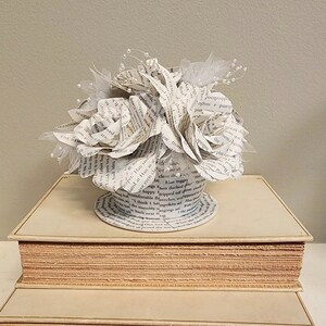 May include: A white paper flower bouquet made from book pages sits on top of a stack of three books. The bouquet is decorated with white tulle and pearl beads.