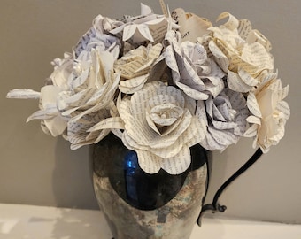 May include: A bouquet of paper roses made from book pages in a silver vase with three feet. The roses are white and cream colored.