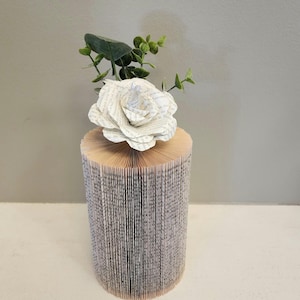 May include: A white paper rose with green leaves sits on top of a book vase made from pages of a book. The vase is cylindrical and has a white background.