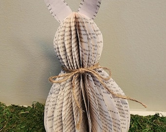 Puede incluir: Una escultura de conejo de papel blanco hecha con las páginas de un libro. El conejo tiene orejas largas y está atado con una cuerda alrededor del medio. El conejo está sentado en un lecho de musgo verde.