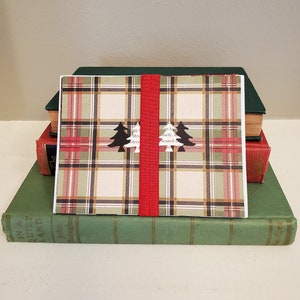 May include: A red and green plaid card with two black Christmas trees on it. The card is tied with a red ribbon and is sitting on top of a stack of books.