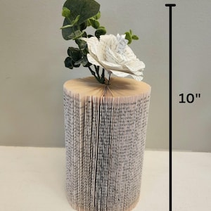 Puede incluir: Un arreglo de flores de papel blanco en un jarr&oacute;n de libro. El jarr&oacute;n est&aacute; hecho de un libro que se ha doblado en forma de cilindro. El jarr&oacute;n mide 10 pulgadas de alto y 4,5 pulgadas de ancho.