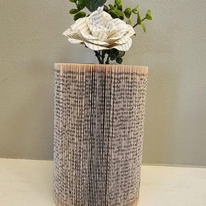 Puede incluir: Una flor de papel blanca con hojas verdes se coloca en un jarr&oacute;n hecho de un libro que se ha doblado en forma de cilindro.