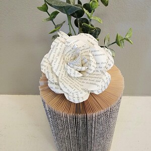 Puede incluir: Una rosa blanca de papel hecha con las p&aacute;ginas de un libro se coloca sobre un jarr&oacute;n de libro. El jarr&oacute;n est&aacute; hecho de un libro que se ha doblado en forma de cilindro. La rosa est&aacute; rodeada de hojas verdes de eucalipto.