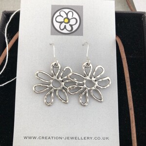 Op de afbeelding: Zilveren bloemvormige oorbellen met een gehamerde textuur, gepresenteerd op een grijze kaart. De oorbellen hebben een haaksluiting. De kaart toont een bloemlogo en het websiteadres: creation-jewellery.co.uk.