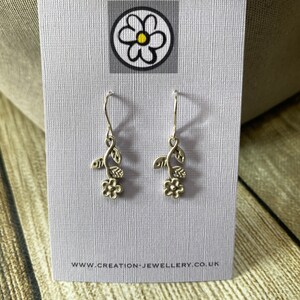 Puede incluir: Pendientes colgantes florales plateados. Cada pendiente presenta un pequeño diseño de flor y hojas. Los pendientes se exhiben en una tarjeta de color púrpura claro con un logotipo y una dirección de sitio web.