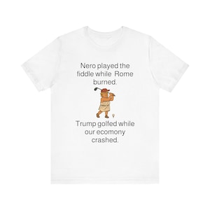 Puede incluir: Camiseta blanca con una caricatura política de un hombre jugando al golf. El texto dice "Nerón tocó el violín mientras Roma ardía. Trump jugó al golf mientras nuestra economía se derrumbaba."