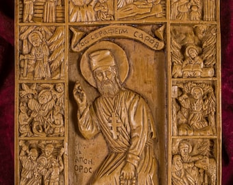 May include: A wooden icon depicting Saint Seraphim of Sarov, a Russian Orthodox saint. The icon is carved in relief and features multiple scenes from his life. The central image shows Saint Seraphim standing with a cross and rosary beads. The icon is framed in a wooden border.