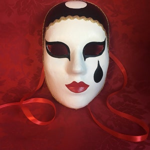 Pode incluir: Uma máscara de carnaval branca com detalhes em preto e dourado. A máscara tem um design de lágrima no lado direito e um círculo preto na parte superior. A máscara está sobre um fundo vermelho.