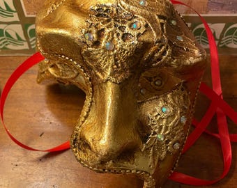 May include: A gold masquerade mask with intricate details and iridescent rhinestones. The mask is tied with a red ribbon.