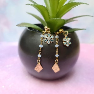 Op de afbeelding: Een paar gouden oorhangers met blauwe sneeuwvlokcharms en een gouden ruitvormige charme aan de onderkant. De oorhangers hangen aan een potplant.