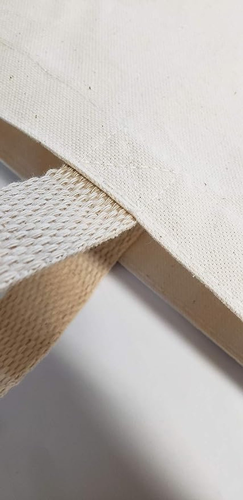 May include: Close-up of a white canvas tote bag with a natural cotton webbing strap. The strap is attached to the bag with a double stitch.