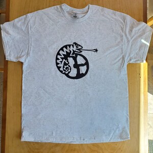 K&ouml;nnte beinhalten: Ein hellgraues T-Shirt mit einem schwarzen Motiv eines Cham&auml;leons, dessen Schwanz um einen Kreis gewickelt ist. Das Cham&auml;leon bl&auml;st einen Luftstrom aus seinem Maul.