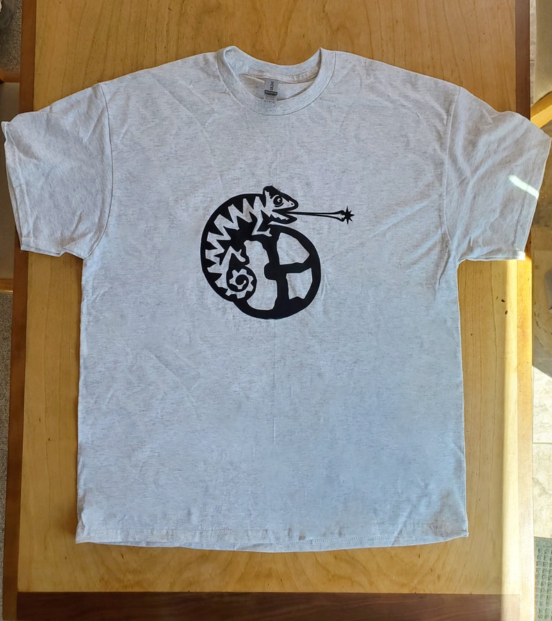 K&ouml;nnte beinhalten: Ein hellgraues T-Shirt mit einem schwarzen Motiv eines Cham&auml;leons, dessen Schwanz um einen Kreis gewickelt ist. Das Cham&auml;leon bl&auml;st einen Luftstrom aus seinem Maul.