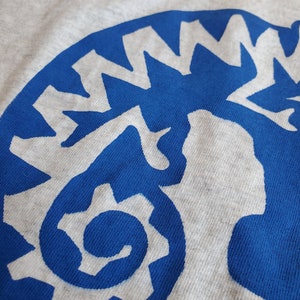 K&ouml;nnte beinhalten: Nahaufnahme eines blauen und wei&szlig;en Grafikdesigns auf einem grauen T-Shirt. Das Design ist eine stilisierte Darstellung eines Zahnrads mit einer Spirale im Inneren.