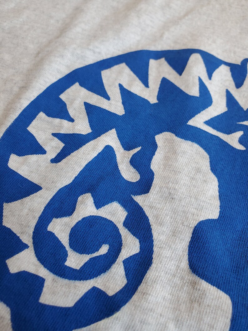 K&ouml;nnte beinhalten: Nahaufnahme eines blauen und wei&szlig;en Grafikdesigns auf einem grauen T-Shirt. Das Design ist eine stilisierte Darstellung eines Zahnrads mit einer Spirale im Inneren.