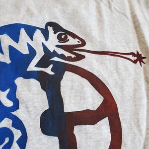 K&ouml;nnte beinhalten: Ein graues T-Shirt mit einem blauen und roten Cham&auml;leon-Motiv. Das Cham&auml;leon blickt nach rechts und hat eine lange Zunge. Das Cham&auml;leon ist schwarz umrandet.