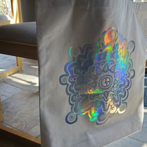 May include: A gray canvas tote bag with a large iridescent holographic mandala design. The mandala features a circular pattern with swirls and dots.