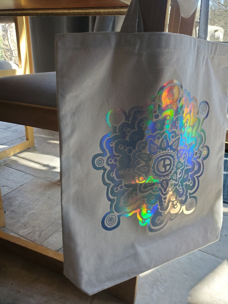 May include: A gray canvas tote bag with a large iridescent holographic mandala design. The mandala features a circular pattern with swirls and dots.