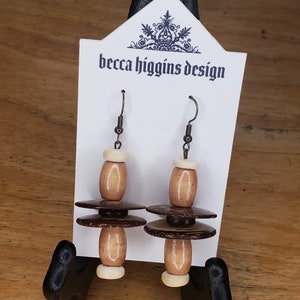 Puede incluir: Un par de pendientes de madera marrón y blanco con un diseño en capas. Los pendientes cuelgan de una tarjeta blanca con el texto "becca higgins design" impreso en ella.