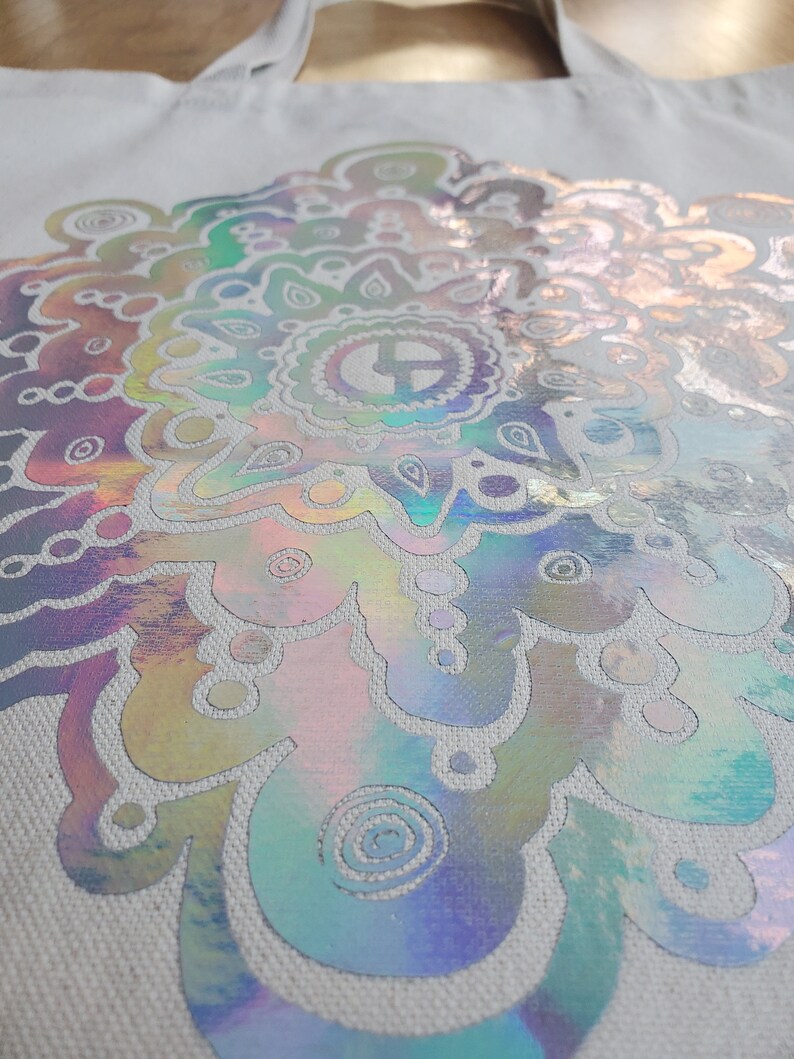 May include: A white canvas tote bag with a large iridescent mandala design. The mandala is a circular pattern with intricate details and a rainbow of colors.