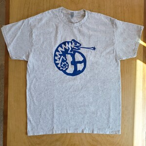 K&ouml;nnte beinhalten: Ein graues T-Shirt mit einem blauen Cham&auml;leon-Motiv. Das Cham&auml;leon ist zu einem Kreis zusammengerollt und hat eine lange Zunge herausgestreckt. Das Motiv ist stilisiert und hat einen Retro-Look.
