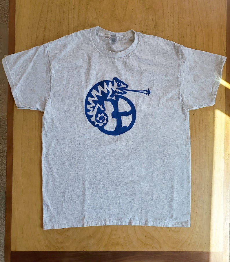 K&ouml;nnte beinhalten: Ein graues T-Shirt mit einem blauen Cham&auml;leon-Motiv. Das Cham&auml;leon ist zu einem Kreis zusammengerollt und hat eine lange Zunge herausgestreckt. Das Motiv ist stilisiert und hat einen Retro-Look.