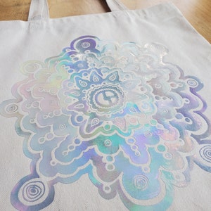 May include: A white canvas tote bag with a large iridescent silver mandala design. The mandala features a swirling pattern of circles and dots.
