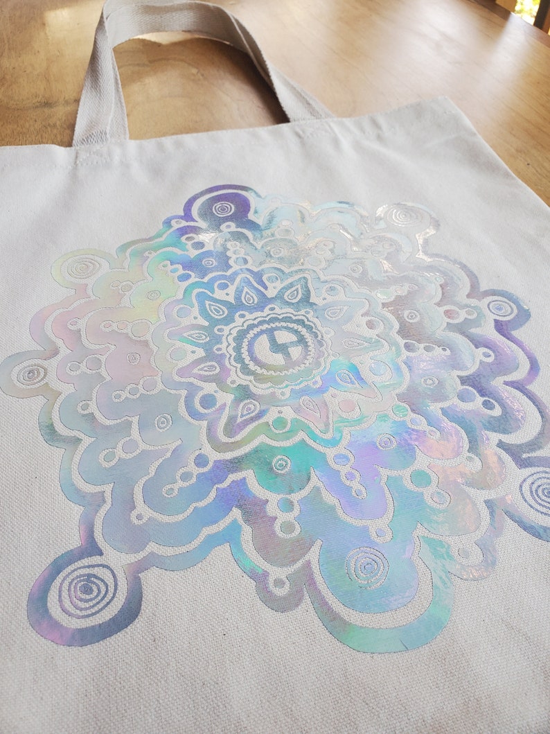 May include: A white canvas tote bag with a large iridescent silver mandala design. The mandala features a swirling pattern of circles and dots.