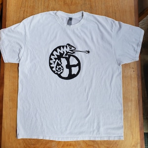 K&ouml;nnte beinhalten: Wei&szlig;es T-Shirt mit einem schwarzen Grafikdruck eines Cham&auml;leons mit einem Stern am Schwanz, das sich um einen Kreis windet.